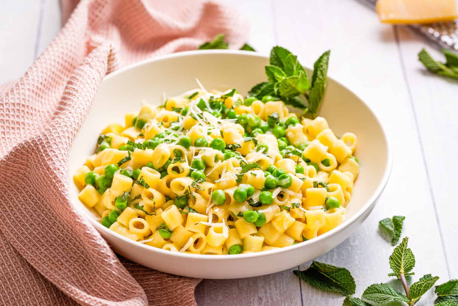 A bowl of short pasta mixed with green peas, grated cheese, and fresh mint, placed on a white table with a pink cloth nearby.