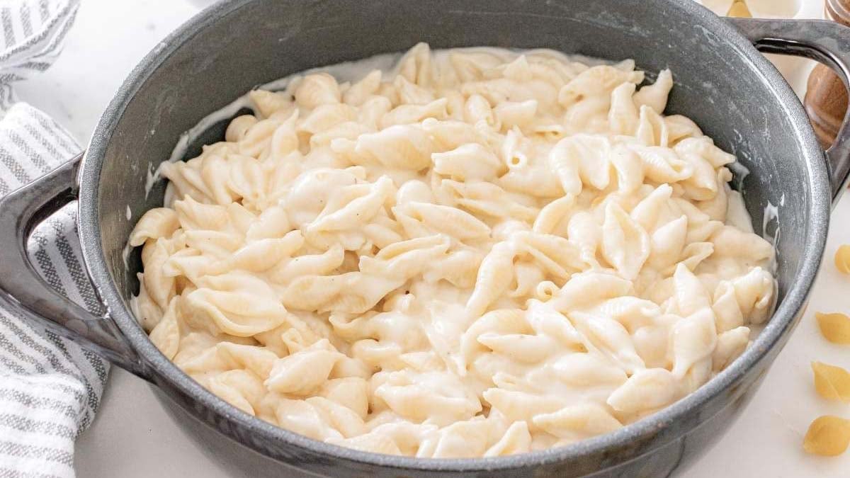 A pot filled with shell pasta coated in creamy white cheese sauce sits on a counter next to a striped towel.