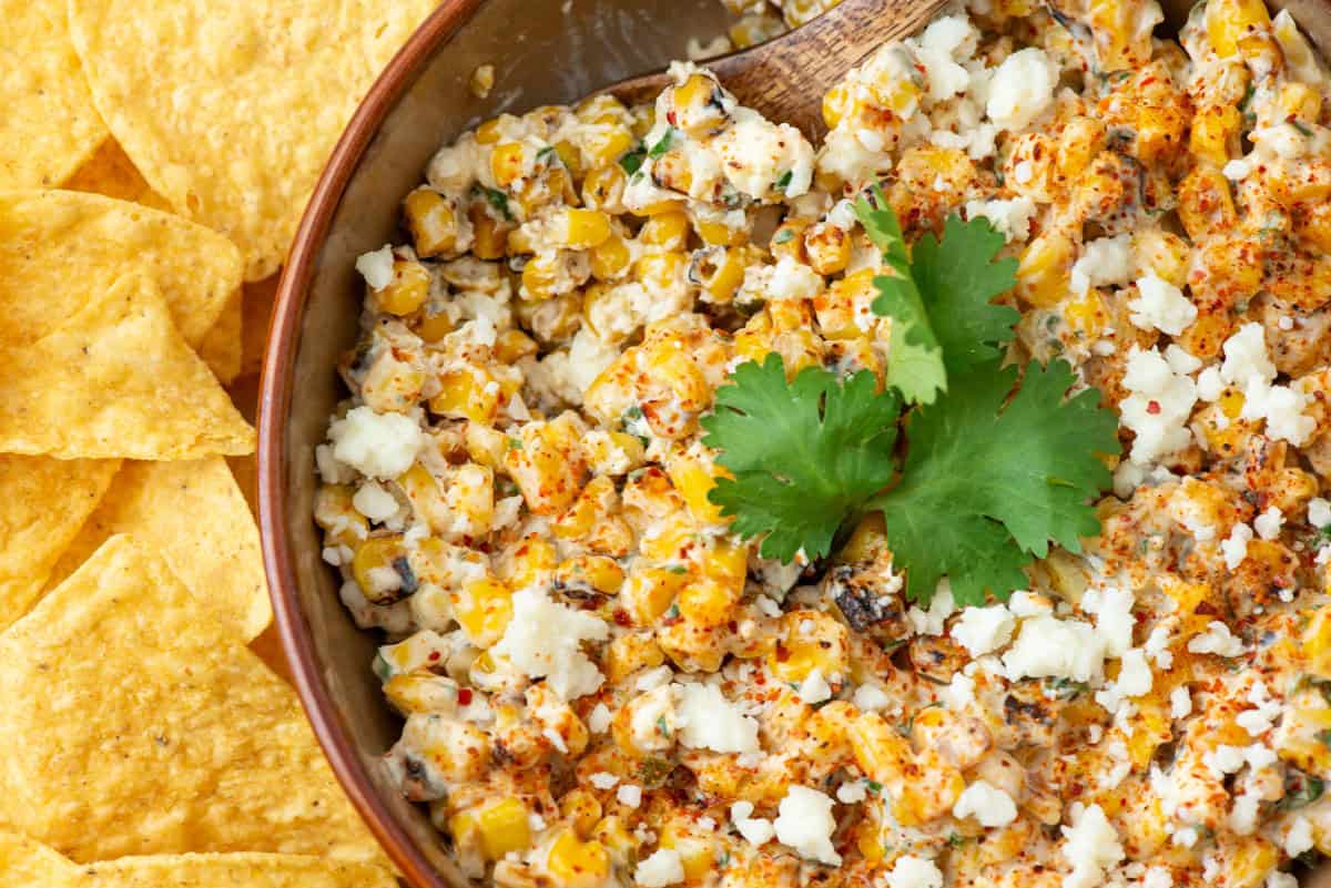 A bowl of Mexican street corn dip topped with crumbled cheese, cilantro, and spices, with tortilla chips on the side.