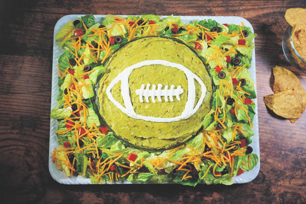 A platter of layered dip arranged and decorated to look like a football field, with guacamole, sour cream, lettuce, shredded carrots, tomatoes, and olives. Tortilla chips are on the side.