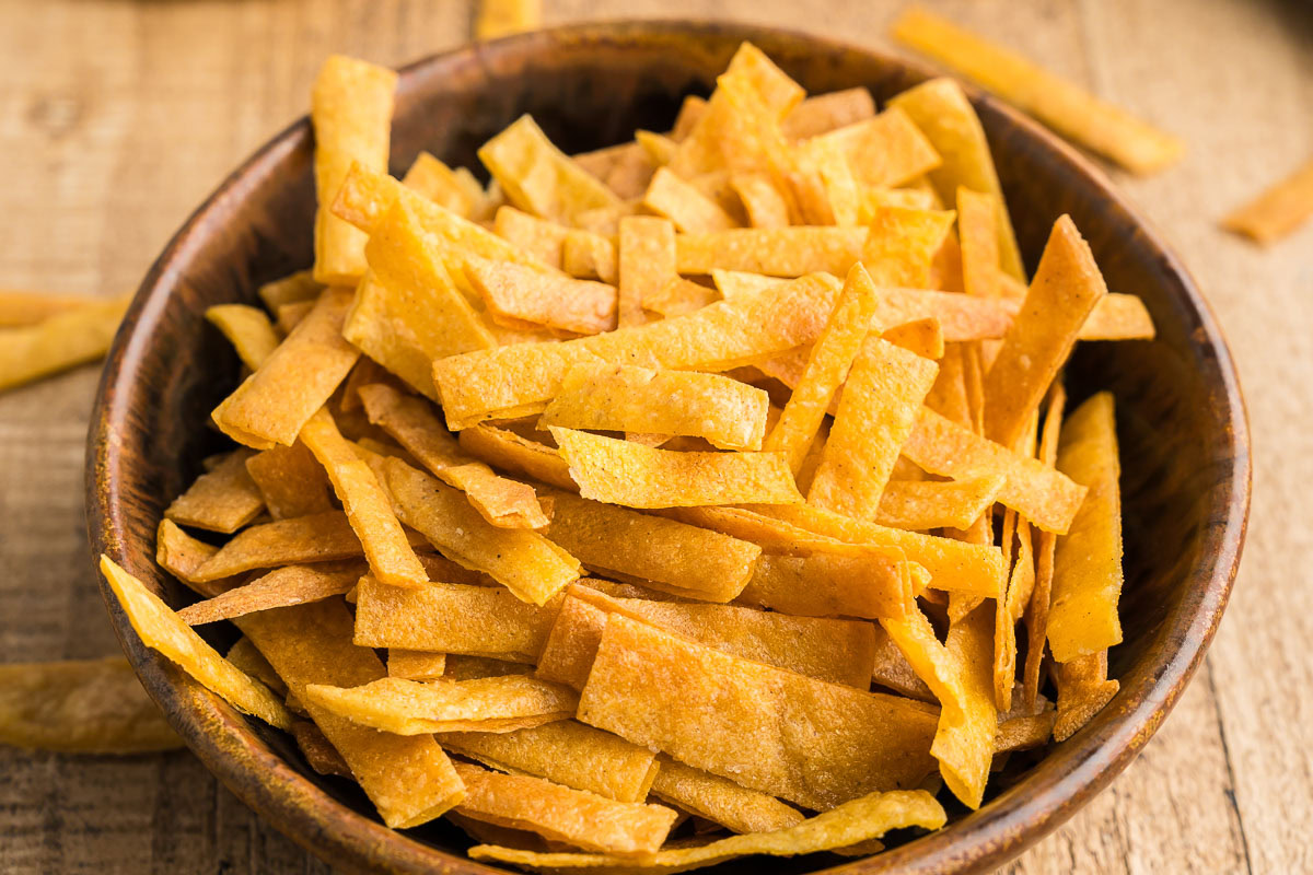 A brown bowl filled with crispy, golden, thinly sliced tortilla strips on a wooden surface.