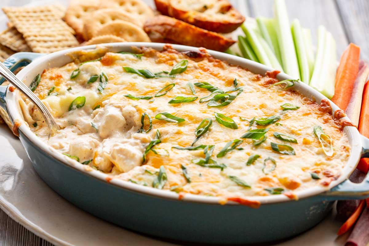 A baked casserole dish filled with cheesy dip, garnished with green onions, served with crackers, toasted bread, and fresh celery and carrot sticks on the side.