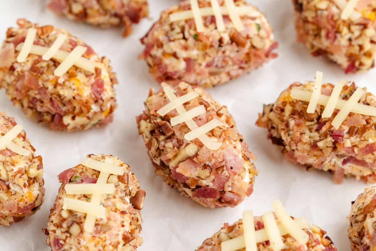 Mini football-shaped cheese balls coated in nuts and bacon, topped with strips of cheese to resemble football laces, arranged on a sheet of parchment paper.