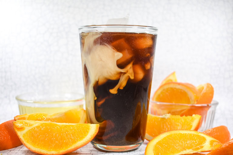 A glass of iced coffee with cream swirling inside, surrounded by orange slices and small glass bowls containing orange segments.