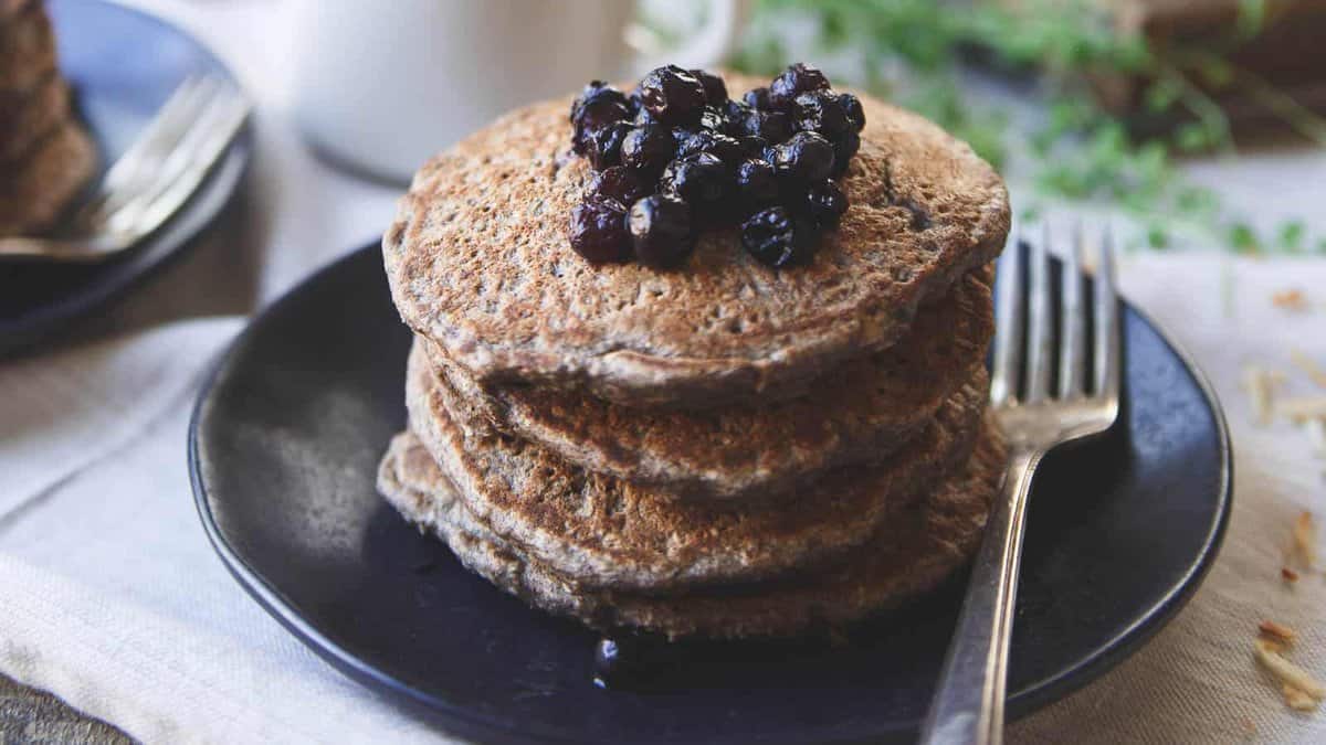 A stack of pancakes topped with blueberries on a dark plate with a fork beside them.
