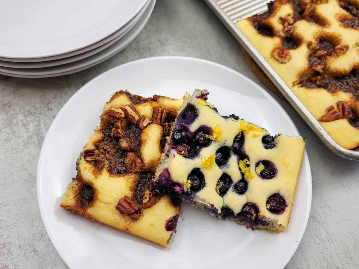 Two slices of cake on a white plate: one with blueberries and lemon zest, the other with pecans and cinnamon topping. A baking tray and stacked plates are in the background.