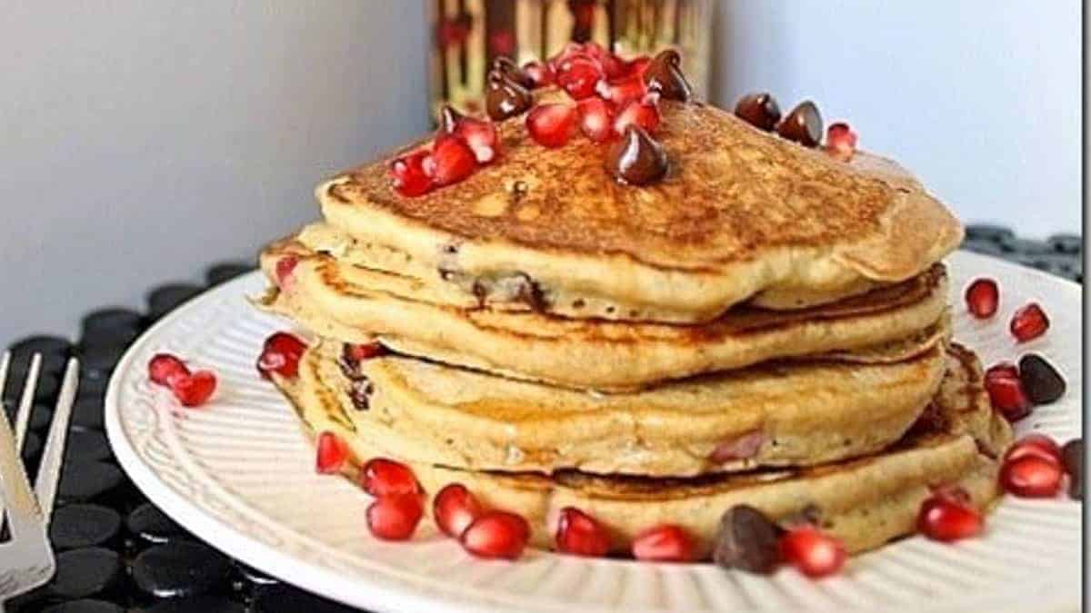 A stack of pancakes topped with pomegranate seeds and chocolate chips sits on a white plate.