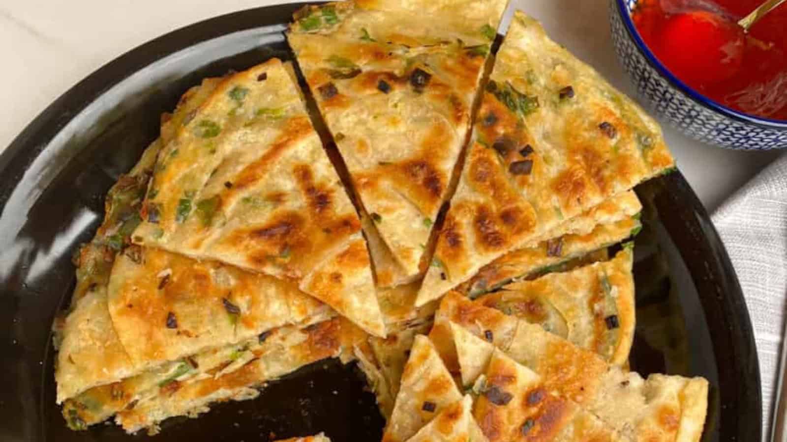 A plate of sliced Chinese scallion pancakes, golden brown with visible green scallions, served next to a small bowl of red dipping sauce.