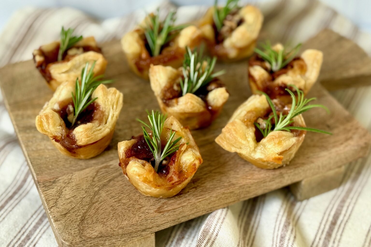 Eight puff pastry tartlets filled with a dark jam and topped with sprigs of fresh rosemary are arranged on a wooden board.