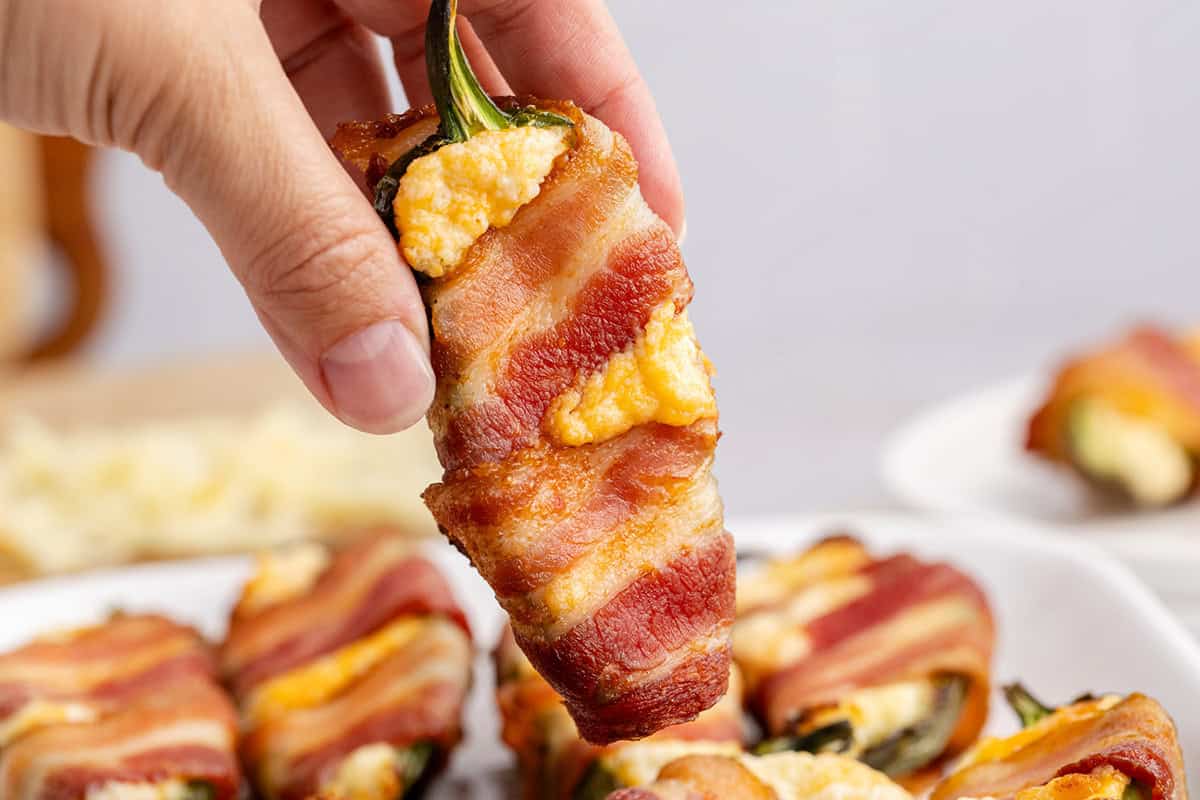 A hand holding a bacon-wrapped jalapeรฑo popper stuffed with cheese, with more poppers on a plate in the background.