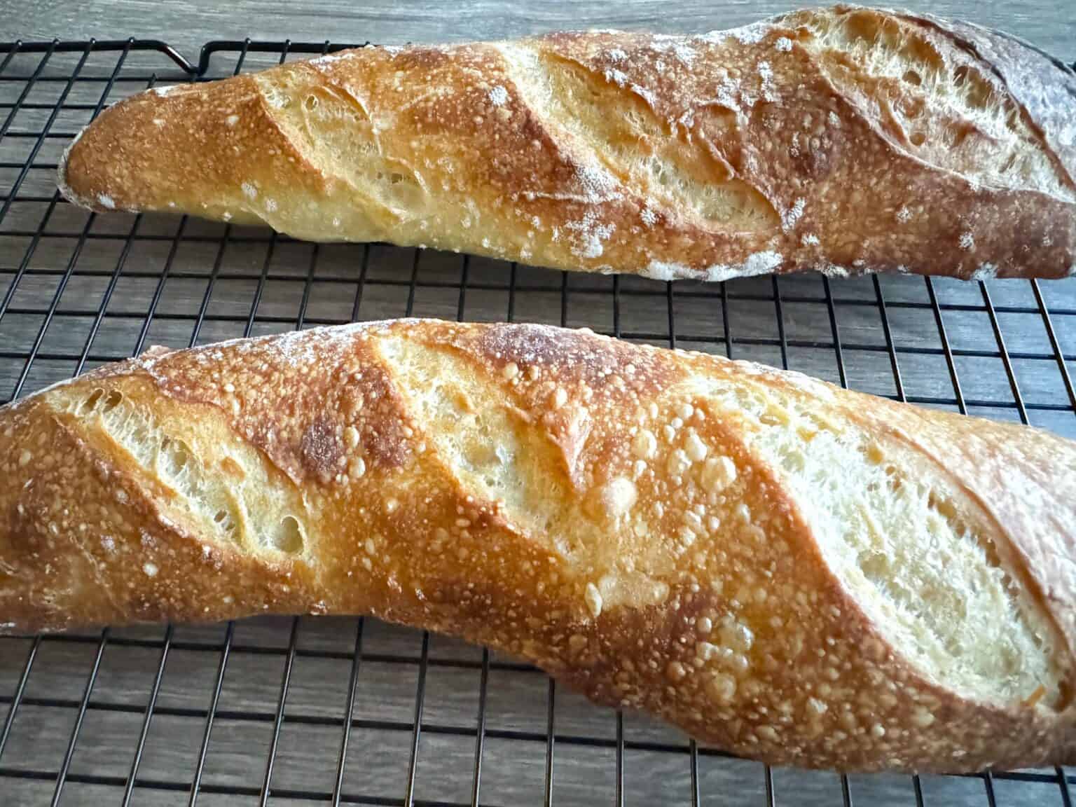 Two freshly baked baguettes with golden, crispy crusts cooling on a black wire rack.