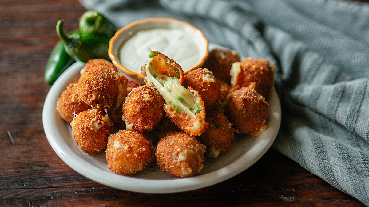 A plate of breaded, fried jalapeรฑo poppers with a bowl of dipping sauce and whole jalapeรฑos, set on a wooden table with a gray cloth.