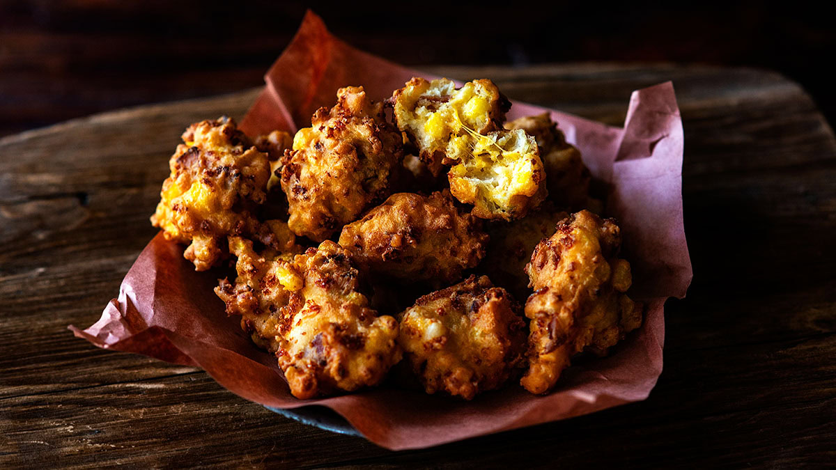 A plate lined with brown parchment paper holds several golden-brown, crispy corn fritters on a wooden surface.