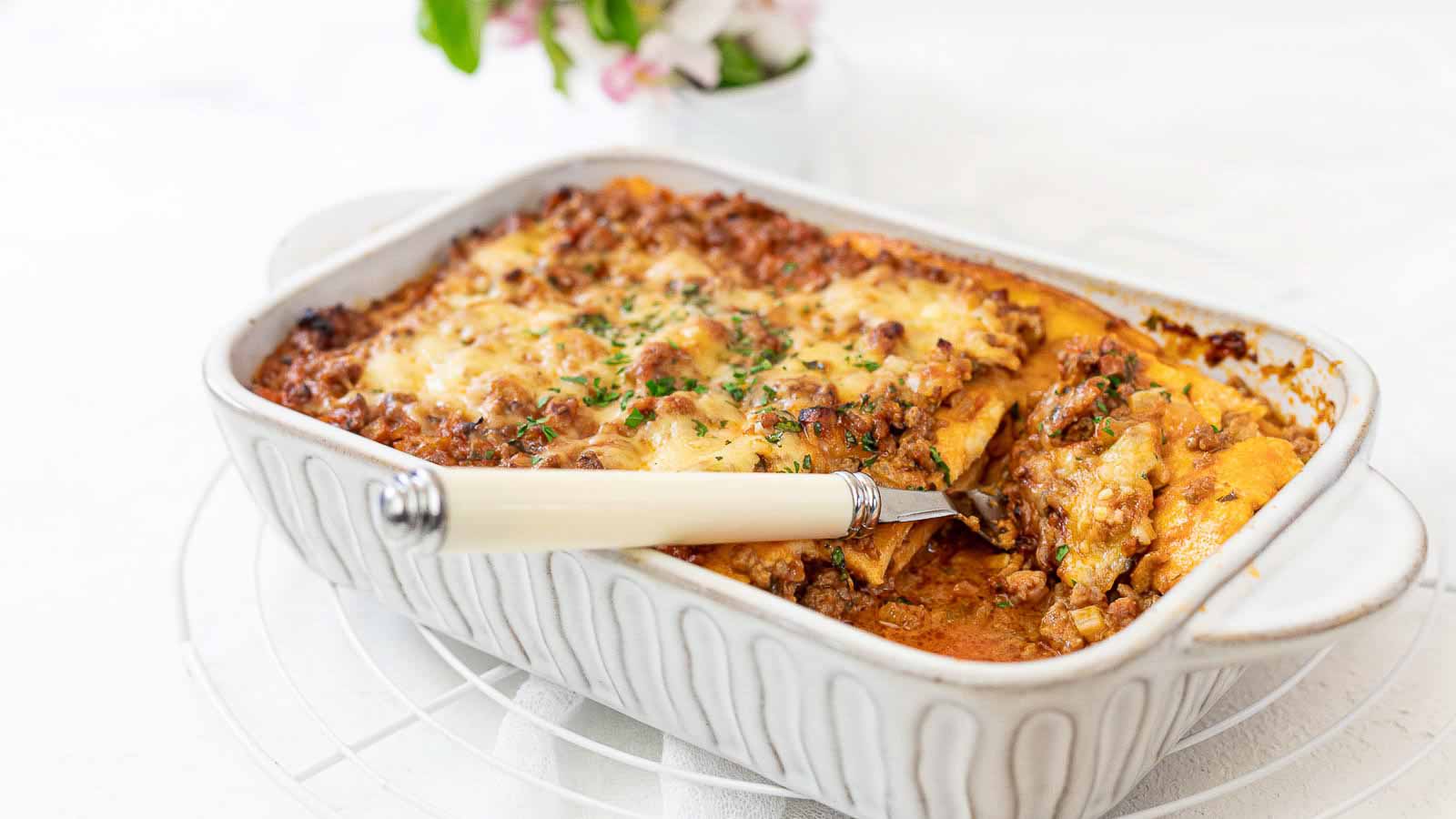 Baked lasagna in a white casserole dish with a serving spoon, showing layers of pasta, meat sauce, and melted cheese.