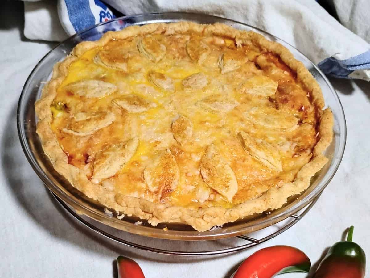 A baked pie with a golden crust and decorative pastry leaves on top, displayed in a glass pie dish on a white surface with red and green peppers nearby.