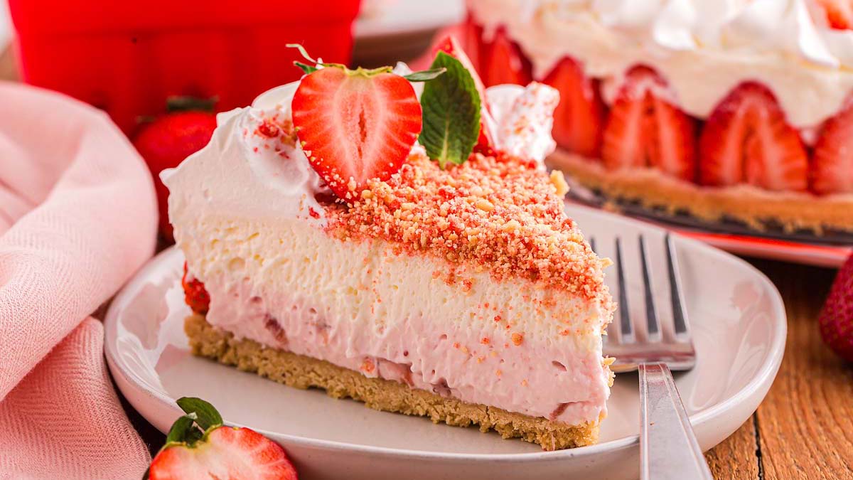 A slice of strawberry cheesecake topped with whipped cream, strawberry halves, and crumb topping, served on a white plate with a fork.