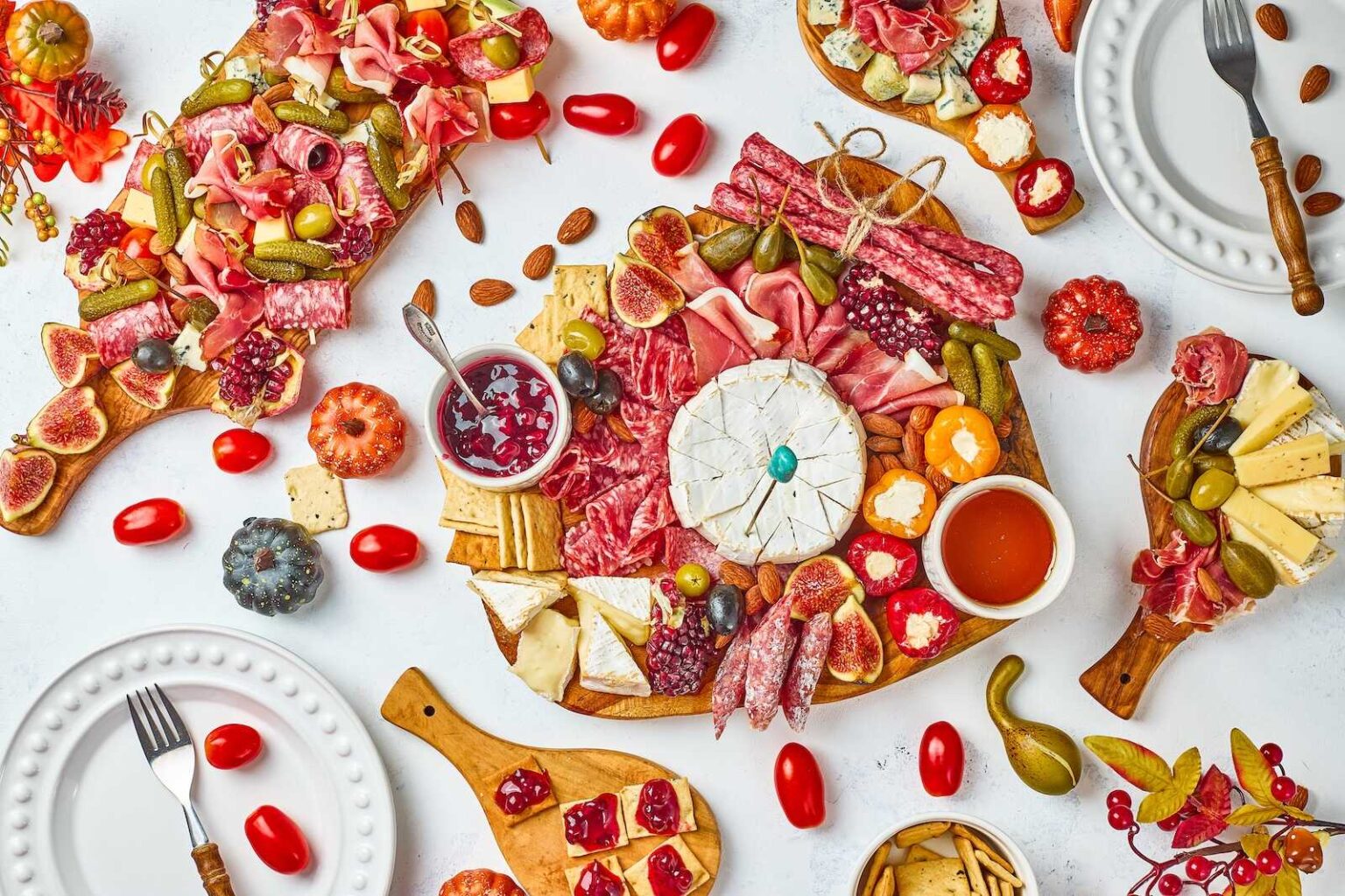 Assorted charcuterie boards with cured meats, cheeses, crackers, fruits, nuts, spreads, and pickles on a white table, surrounded by plates, utensils, and grape tomatoes.