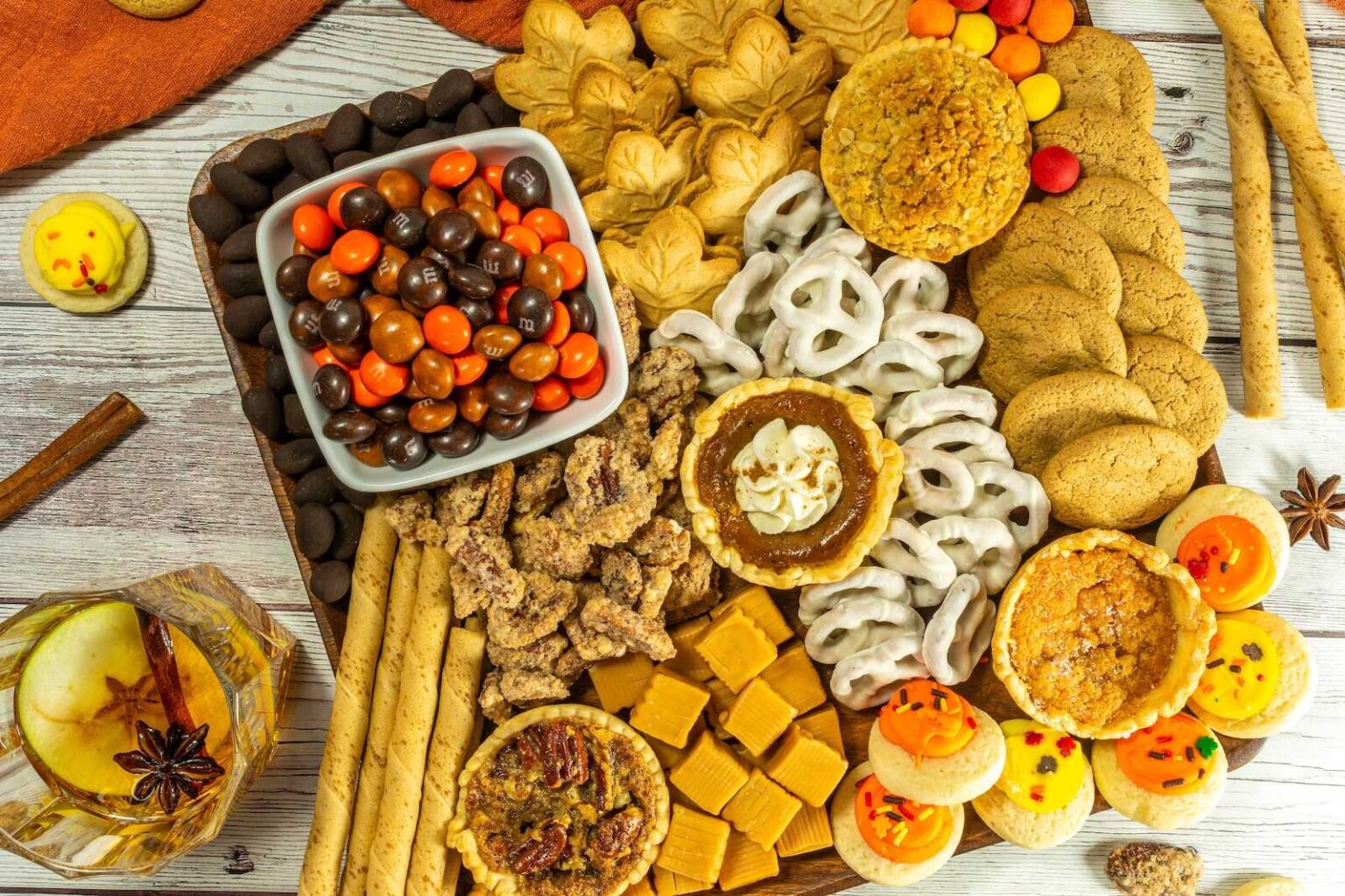 A wooden board with assorted cookies, mini pies, white chocolate pretzels, caramel fudge, chocolate candies, candied nuts, and a glass of apple cider garnished with cinnamon and star anise.