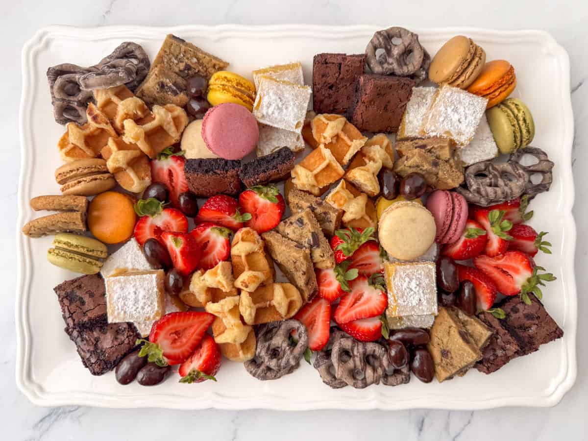 A white platter with assorted desserts, including brownies, strawberries, waffles, macarons, chocolate-covered pretzels, powdered sugar squares, and cookies.