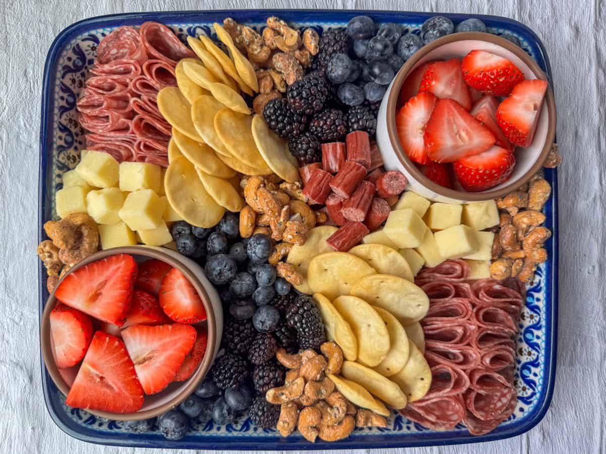 A platter with sliced salami, cheese cubes, round crackers, candied nuts, blueberries, blackberries, dried sausage, and two bowls of sliced strawberries.
