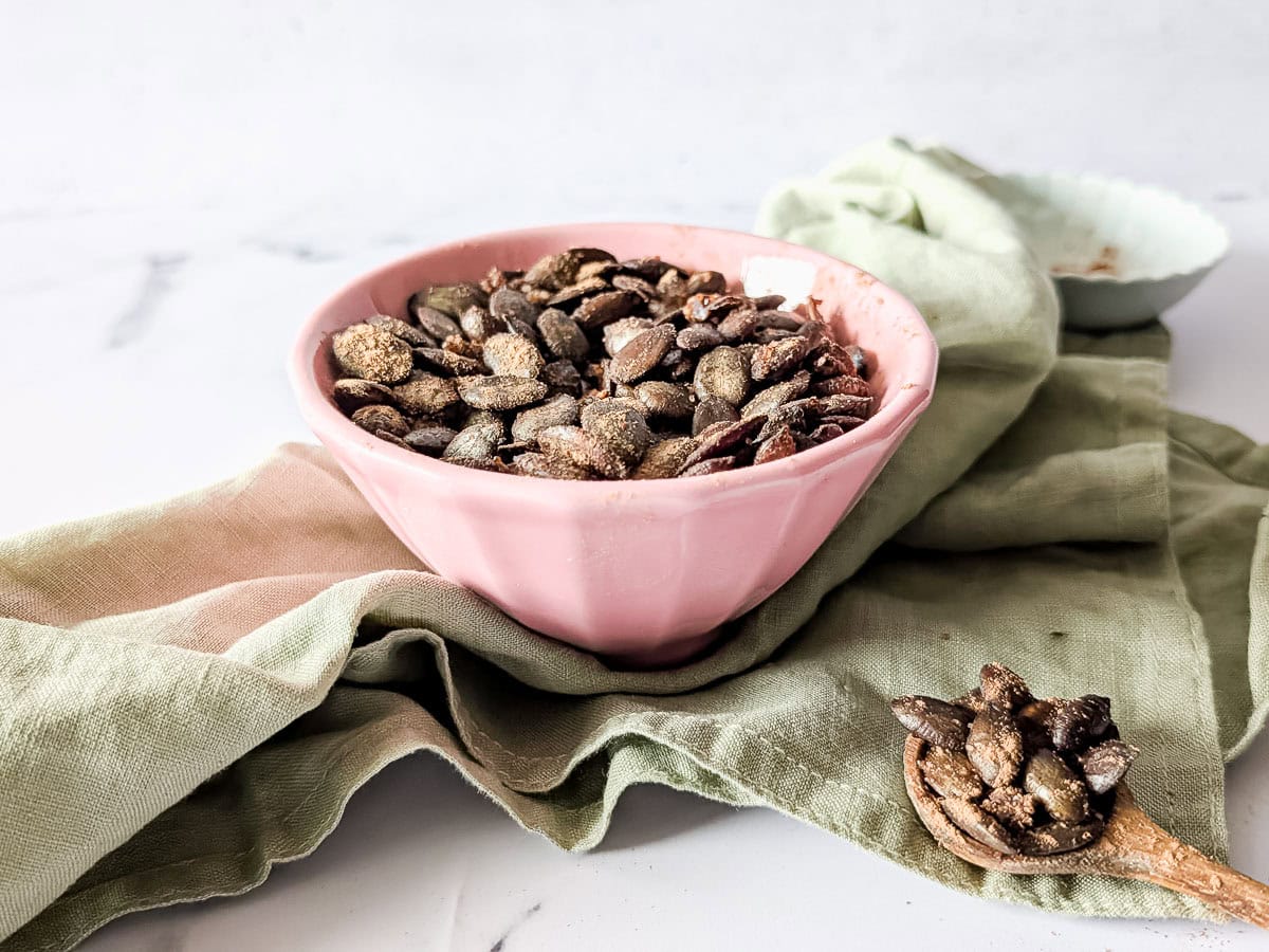 A pink bowl filled with roasted pumpkin seeds sits on a green cloth, with a wooden spoon holding seeds in the foreground.