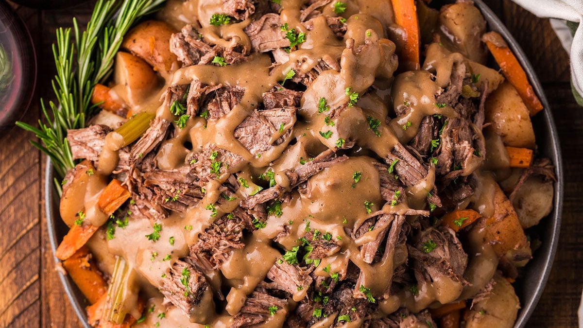 A plate of pot roast topped with brown gravy, surrounded by carrots, potatoes, and garnished with chopped herbs and a sprig of rosemary.