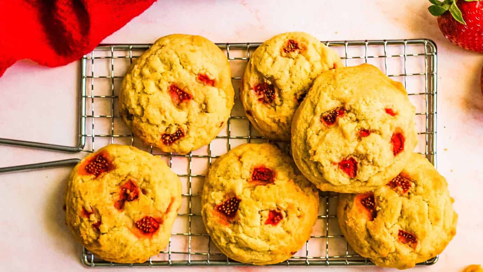 Six strawberry-studded cookies are cooling on a metal rack, with a red cloth and a strawberry partially visible in the corners.
