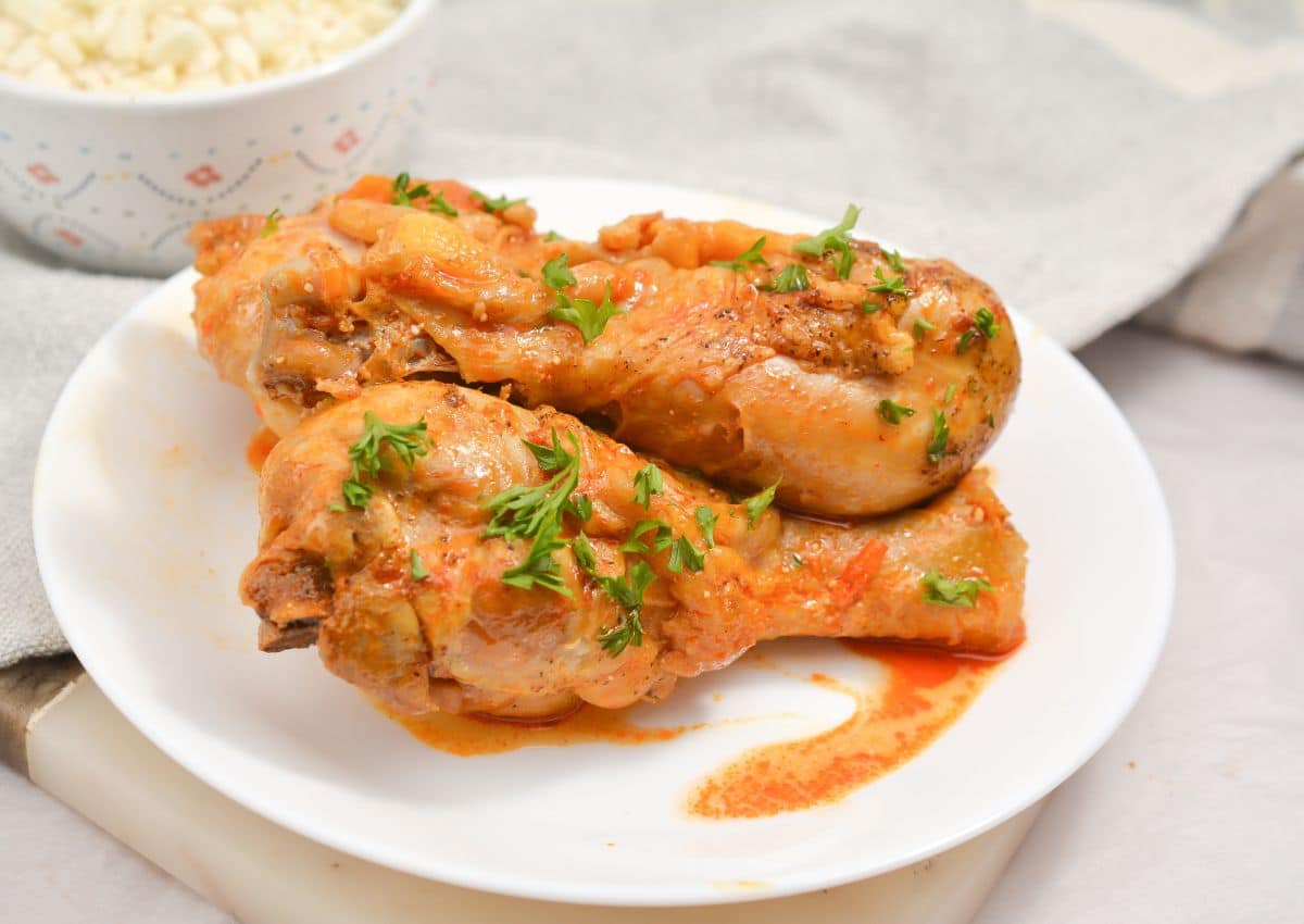 Two cooked chicken drumsticks with sauce and chopped herbs served on a white plate, with a bowl of rice in the background.
