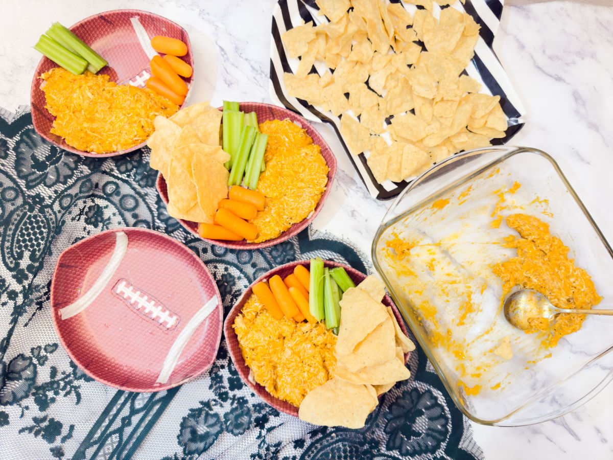 Three football-themed bowls hold dip, celery sticks, baby carrots, and tortilla chips. A larger dish of dip with a spoon and a tray of tortilla chips are also on the table.