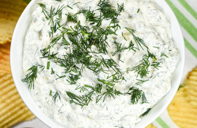 A bowl of creamy dip topped with chopped fresh dill, surrounded by ridged potato chips.