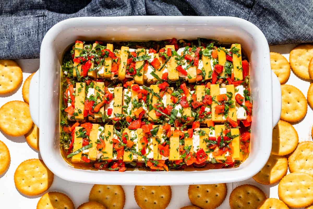 Rectangular dish with layered cubes of cheese, herbs, and chopped red peppers in oil, surrounded by round crackers.