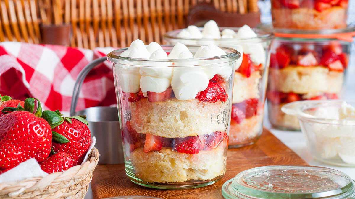 Glass jars filled with layered sponge cake, sliced strawberries, and dollops of whipped cream, with fresh strawberries and a checkered cloth nearby.