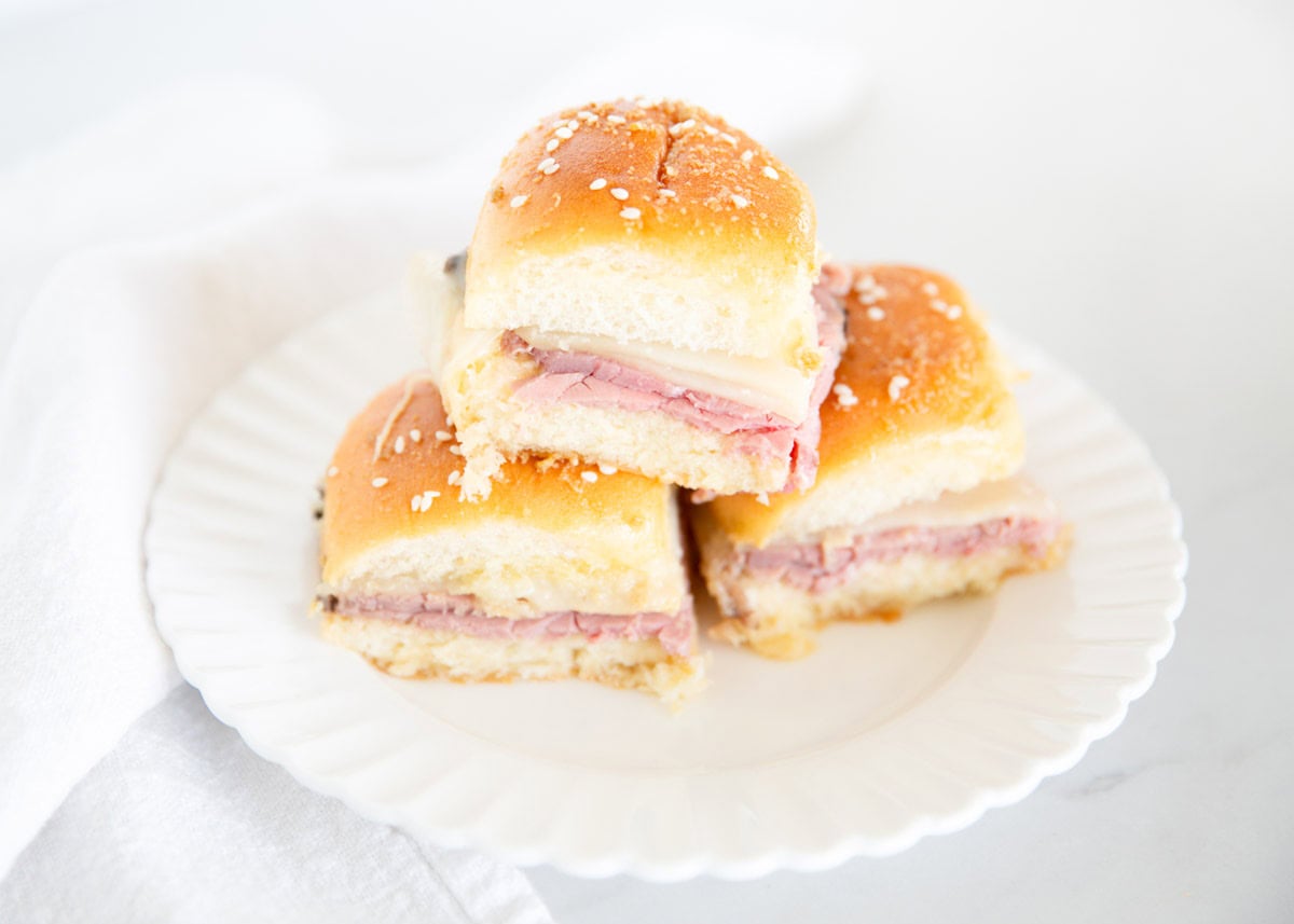 Three ham and cheese sliders on a white plate with a white napkin in the background.