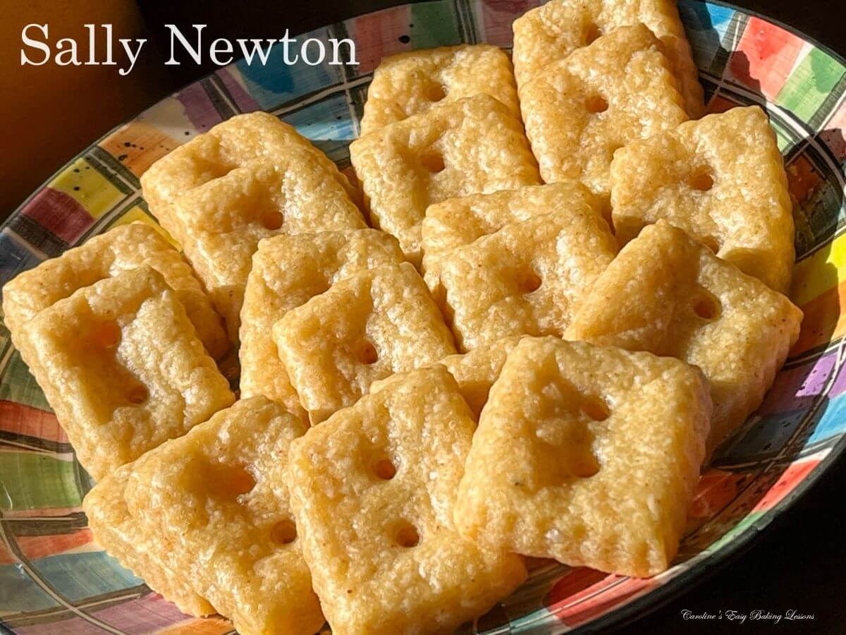 A colorful bowl filled with square, golden crackers that have two holes in the center of each piece. The words Sally Newton are overlaid in the top left corner.