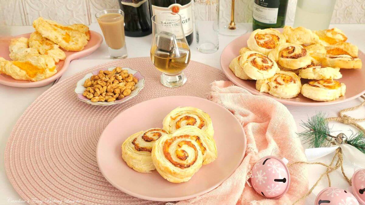 Plates of puff pastry pinwheels, scones, and a dish of peanuts are arranged on a pink placemat with glasses of drinks, wine bottles, and festive ornaments.