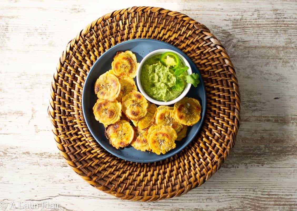 A plate of tostones with a small bowl of guacamole is placed on a round wicker placemat atop a light wooden surface.