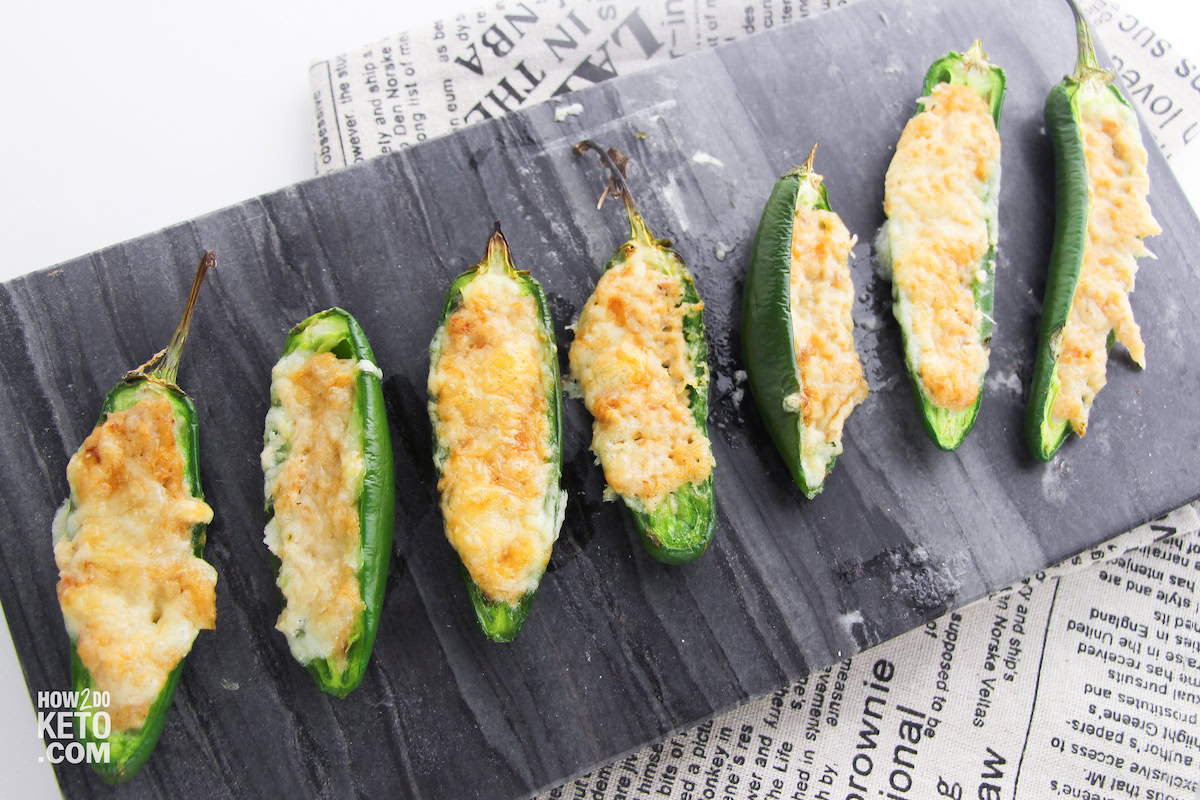 Seven jalapeรฑo halves filled with melted, browned cheese are arranged in a row on a slate serving board placed over newspaper print.