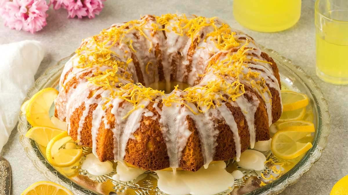 A lemon bundt cake topped with white icing and lemon zest sits on a glass platter, surrounded by lemon slices.