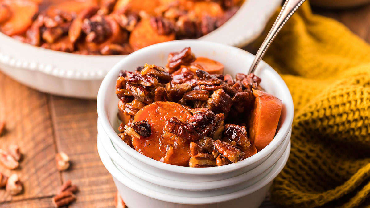 A bowl of candied sweet potatoes topped with pecans, with a spoon in the bowl and a casserole dish in the background.