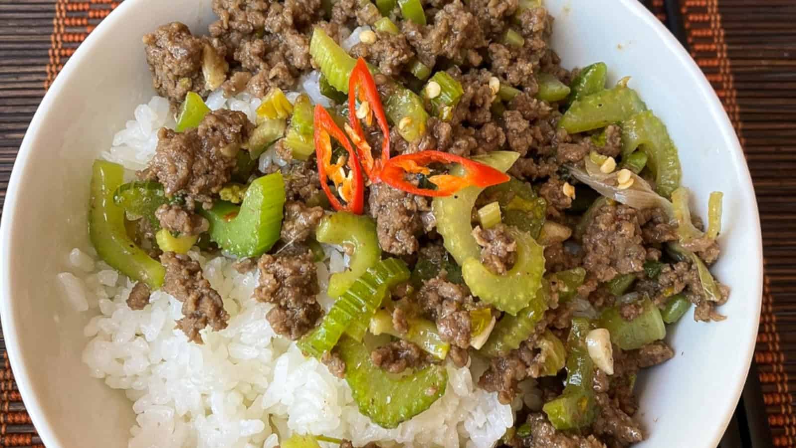 A bowl of white rice topped with stir-fried ground beef, celery slices, chopped vegetables, and garnished with red chili slices.