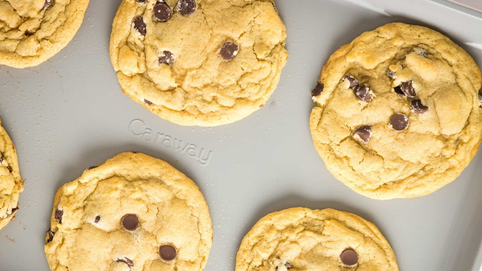 Six chocolate chip cookies on a gray baking sheet, spaced apart, with visible chips on the tops of the cookies.