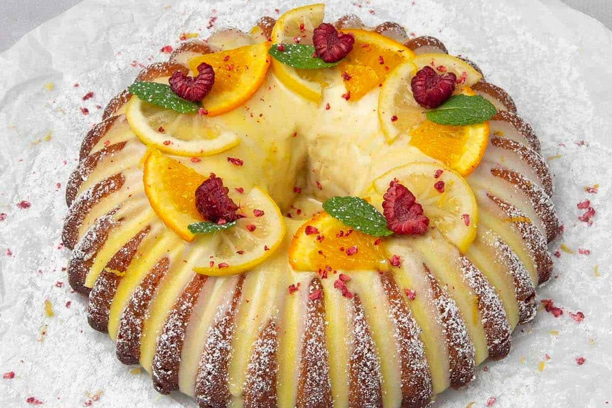 Bundt cake topped with lemon and orange slices, raspberries, mint leaves, and powdered sugar, set on a white surface.