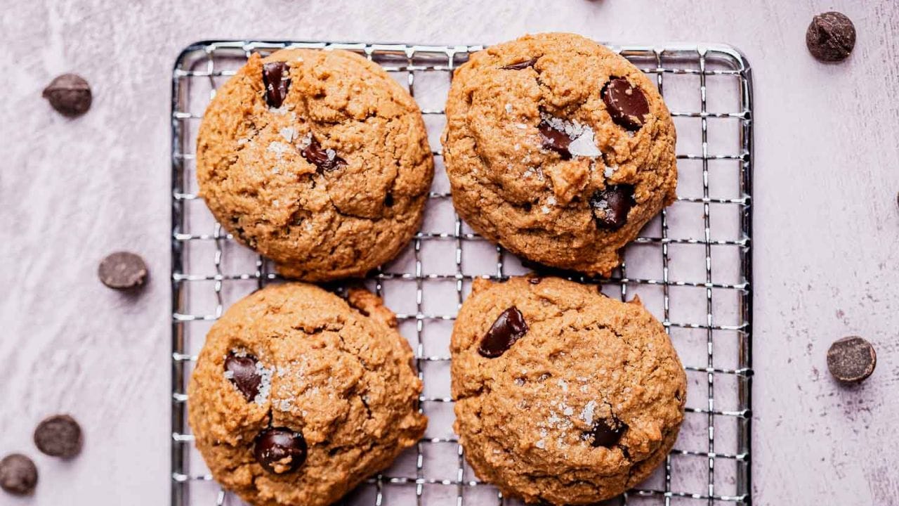 Four chocolate chip cookies on a wire cooling rack with scattered chocolate chips on a light surface.