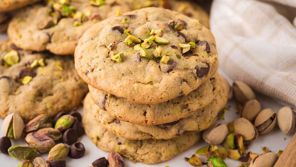 A stack of chocolate chip cookies topped with chopped pistachios, surrounded by whole pistachios and chocolate chips on a table.