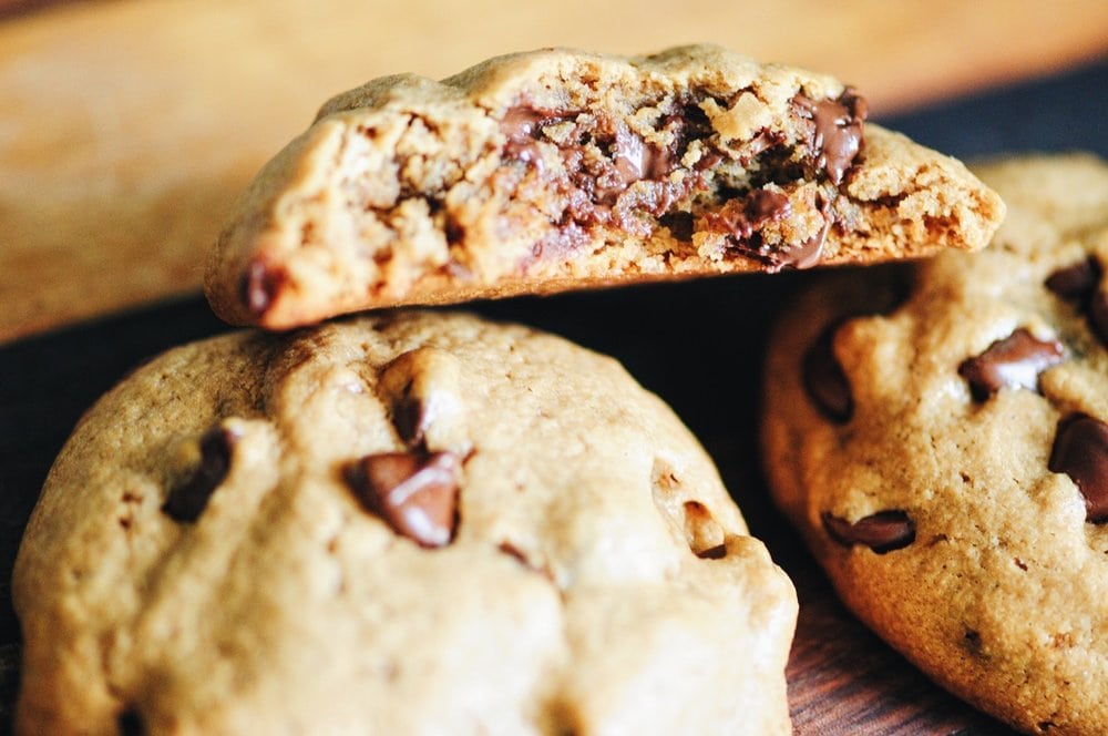 Three chocolate chip cookies on a wooden surface, with one cookie broken in half to show the soft, chocolate-filled interior.