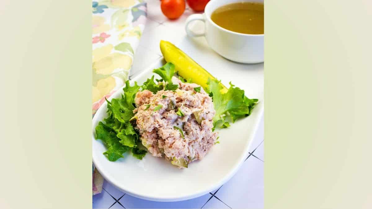 A scoop of tuna salad on lettuce with a pickle spear, served on a white plate next to a cup of soup and a floral napkin.
