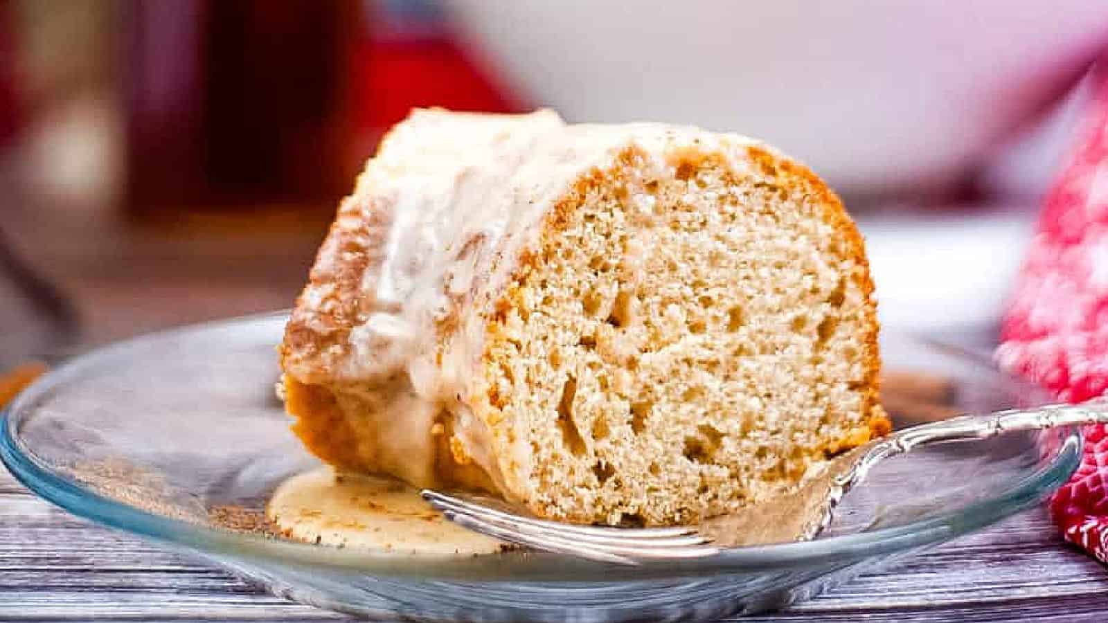 A slice of glazed bundt cake sits on a glass plate with a fork, showing a light, fluffy texture and creamy icing.