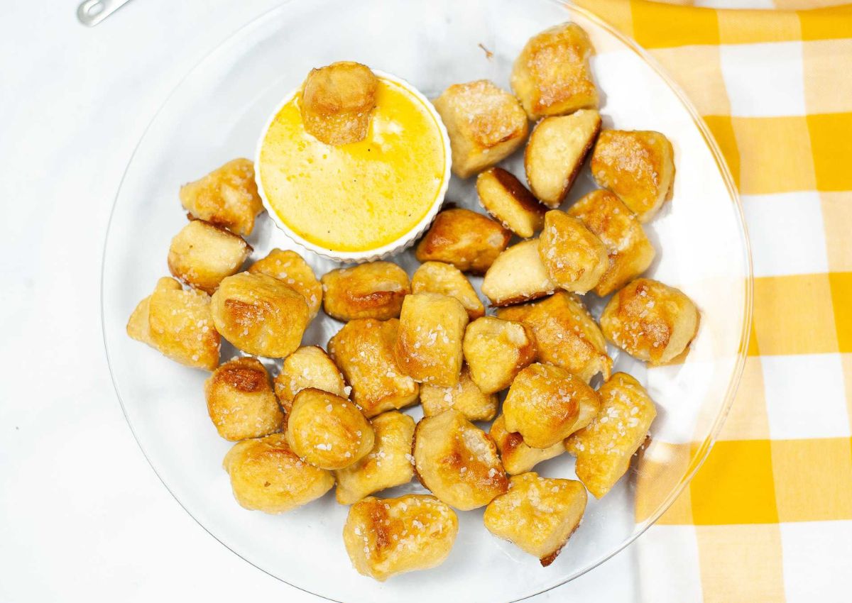 A clear plate of soft pretzel bites with coarse salt, served with a small dish of yellow cheese dip, next to a yellow and white checkered napkin.