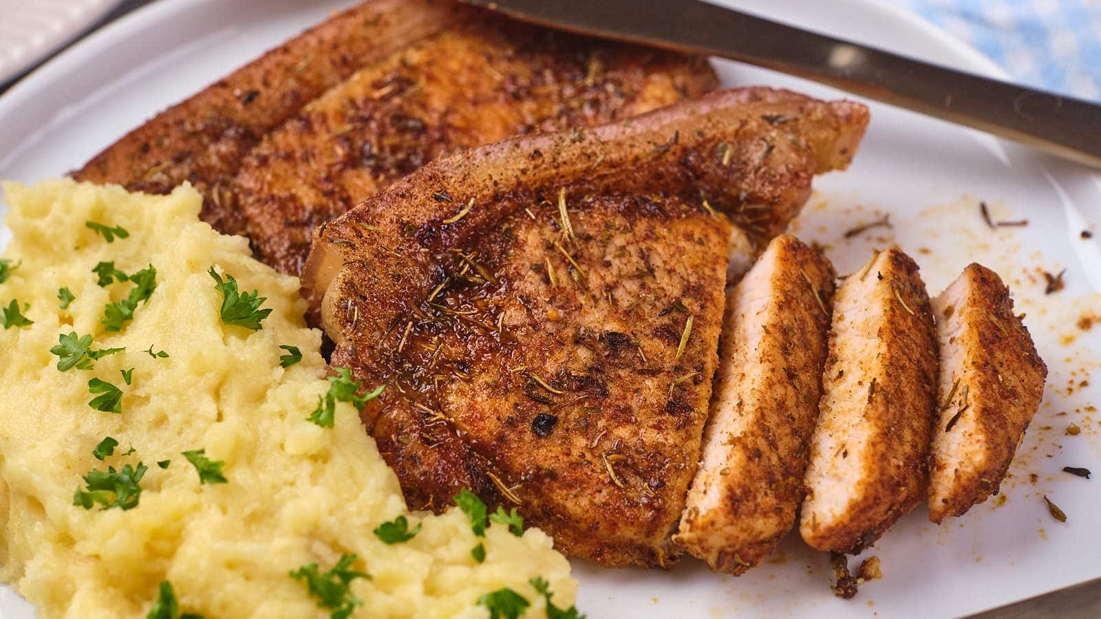 Seasoned pork chop, partially sliced, served next to a portion of mashed potatoes garnished with chopped parsley on a white plate.