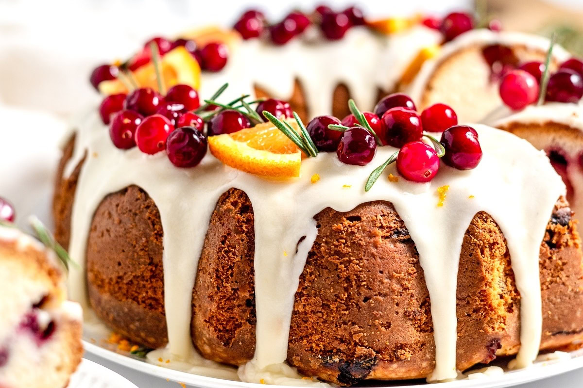 A bundt cake topped with white icing, fresh cranberries, orange slices, and sprigs of rosemary.